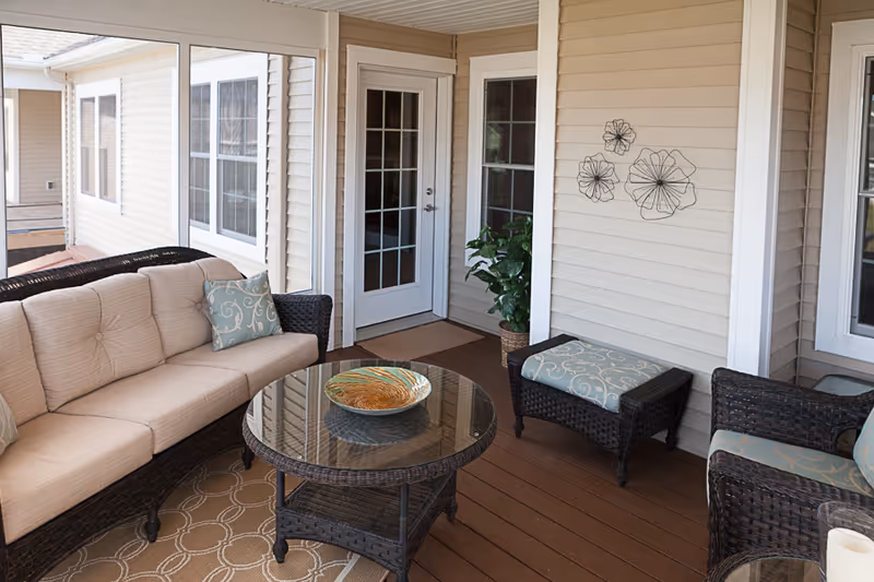 A cozy enclosed patio area with wicker furniture including a beige cushioned sofa, two armchairs, and an ottoman with patterned cushions. A round glass-top coffee table with a decorative bowl sits in the center on a patterned rug. The walls are beige with white trim, decorated with three metal flower wall hangings and a potted plant near a glass door.