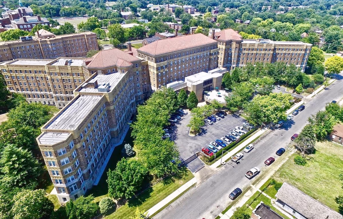 Aerial view of Homer G Phillips Senior Apartments, a large multi-story brick building surrounded by trees and greenery with a parking lot in front and cars parked along the street.