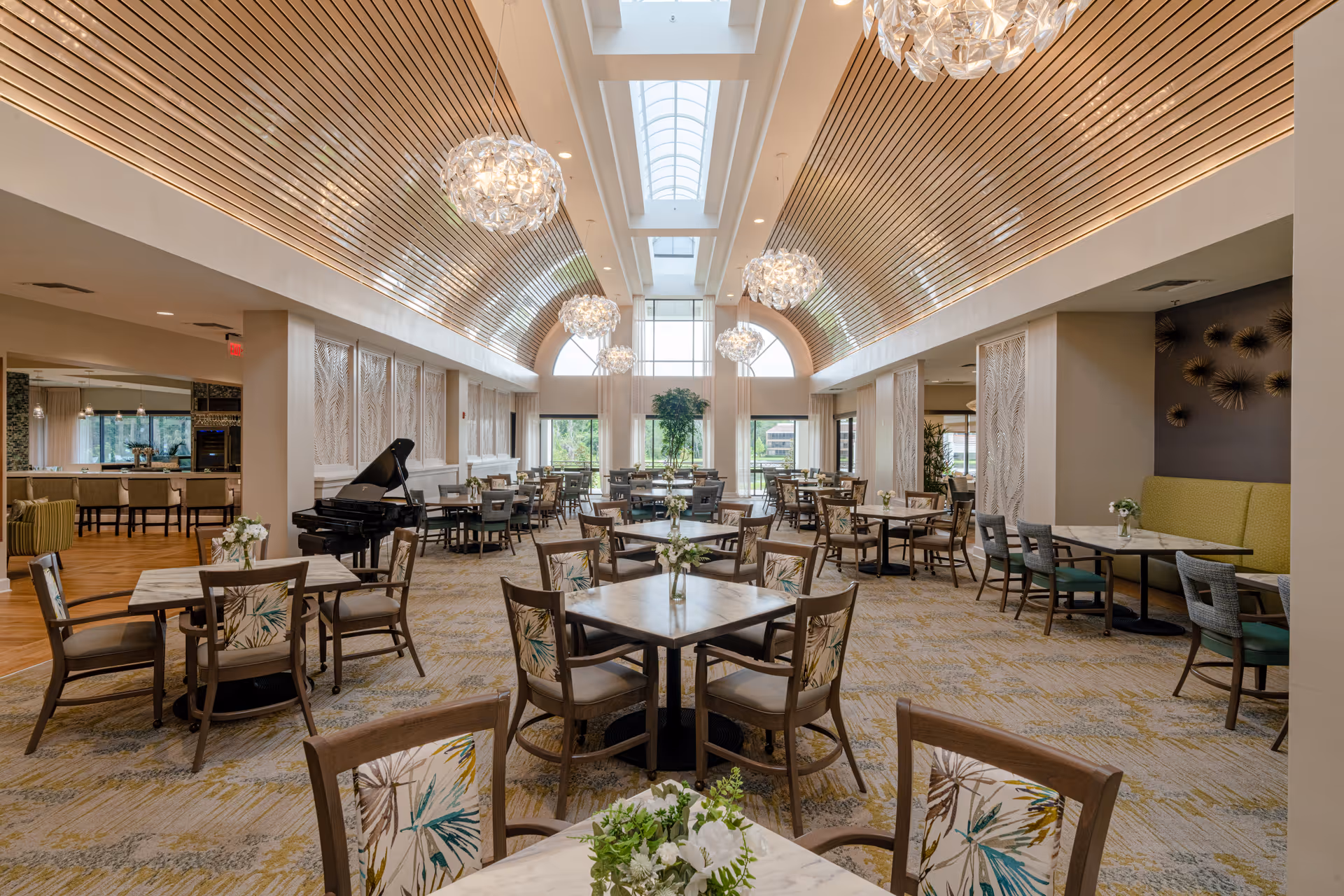 Spacious, elegant dining room with multiple tables and chairs, skylights, chandeliers, and a grand piano.