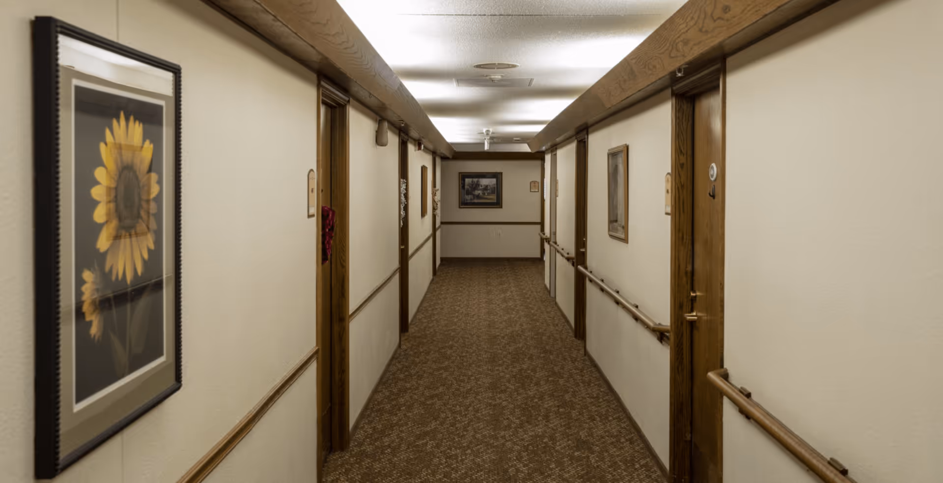 Carpeted interior hallway in an assisted living facility with handrails, framed artwork, and several room doors.