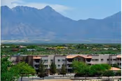 View of a multi-story residential building surrounded by trees with a mountain range in the background under a clear blue sky.