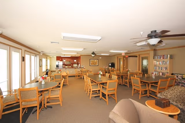 Large communal dining and activity room with multiple tables and chairs, a kitchenette at the back, and a seating area with bookshelves.