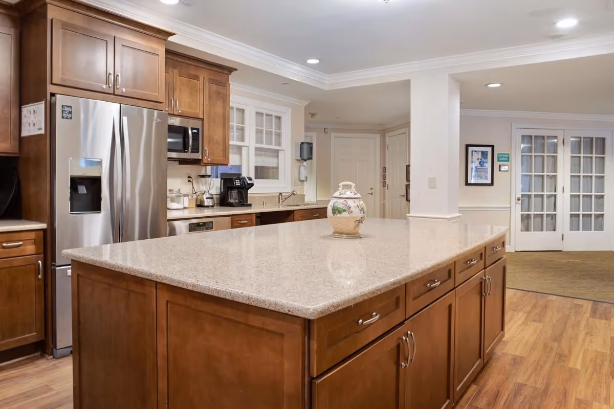 A spacious kitchen area in a senior living facility featuring a large central island with a decorative ceramic vase on top. The kitchen has wooden cabinets, a stainless steel refrigerator with a water dispenser, a microwave, a coffee maker, and a blender. The floor is wooden, and there are white walls with crown molding. In the background, there are double glass doors and a hallway with white doors.