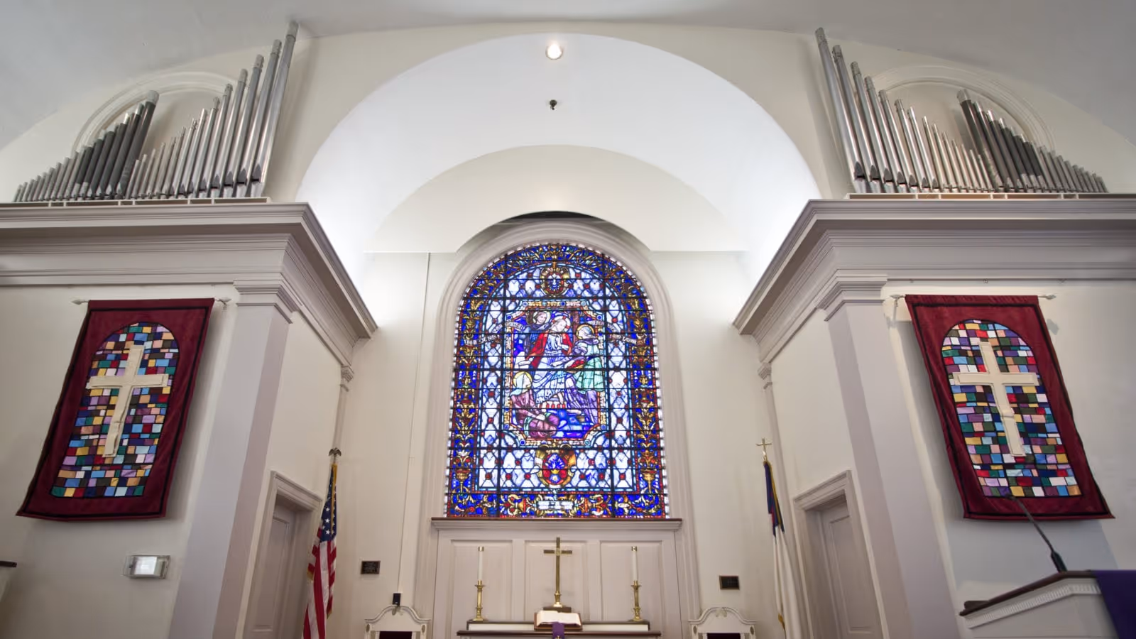 Interior view of a chapel with a large stained glass window depicting a religious scene, two red banners with white crosses on either side, an American flag, a Christian flag, and a pipe organ above the banners.