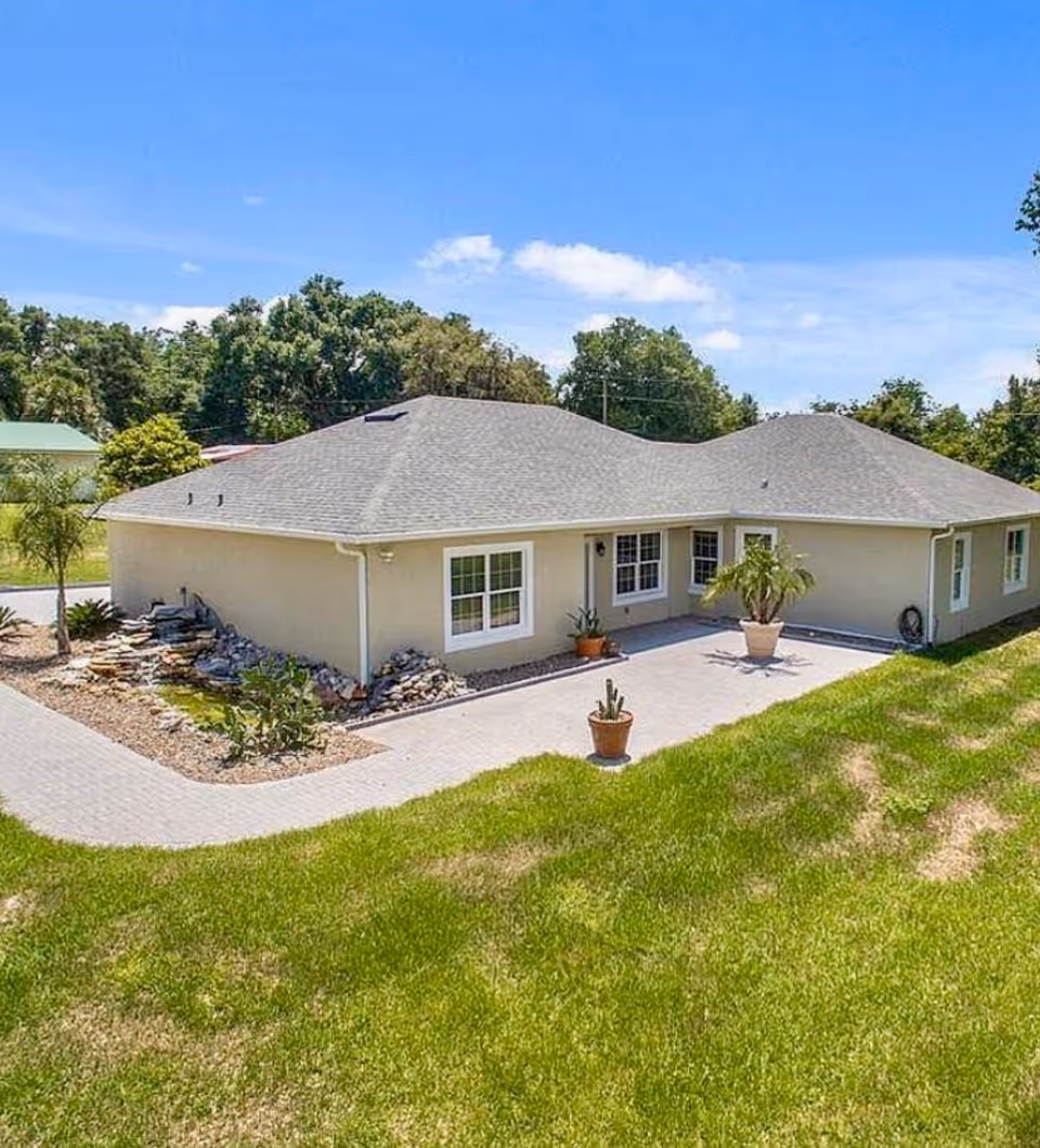 Single-story beige building with a gray shingled roof, surrounded by green grass and trees under a blue sky. There are potted plants on a paved area near the building and a small rock garden with a water feature on the left side.