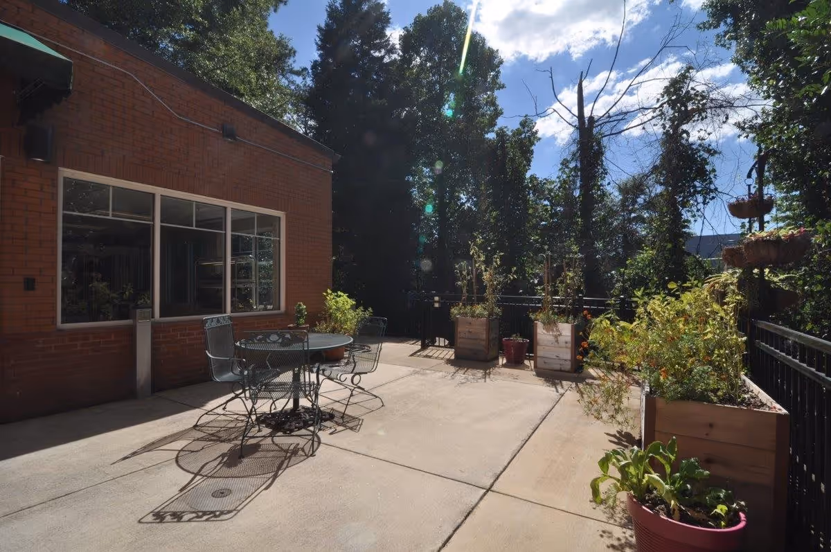 Outdoor patio area with a round metal table and four matching chairs on a concrete surface. The patio is adjacent to a brick building with a large window. Several planters with various plants and flowers are placed along the black metal railing surrounding the patio. Trees and a partly cloudy sky are visible in the background.