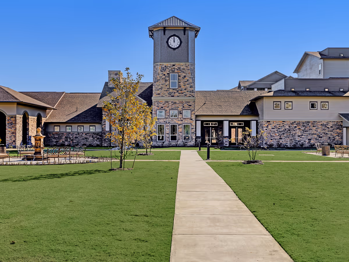 Exterior view of The Lodge at Pine Creek featuring a large building with a clock tower, stone and siding facade, a concrete walkway leading through a green lawn, and clear blue sky.
