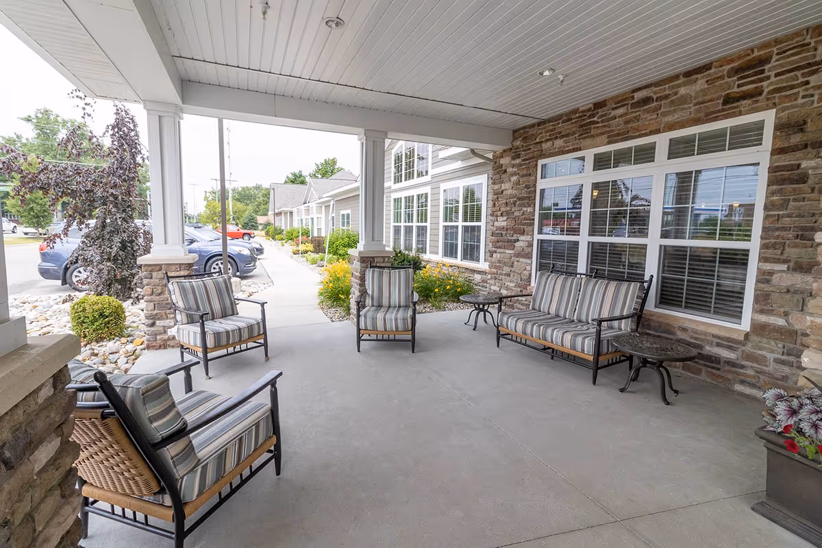 Covered outdoor seating area with cushioned chairs and a loveseat arranged around small tables, adjacent to a stone and siding building with large windows, overlooking a parking lot and landscaped walkway.