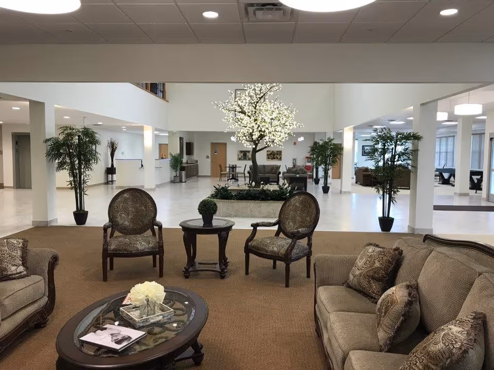 A spacious and well-lit senior living facility common area with comfortable upholstered sofas and armchairs arranged around a glass-top coffee table. In the background, there is a decorative illuminated tree centerpiece surrounded by plants, with additional seating and tables further back. The area has a high ceiling with recessed lighting and white walls, creating an open and welcoming atmosphere.