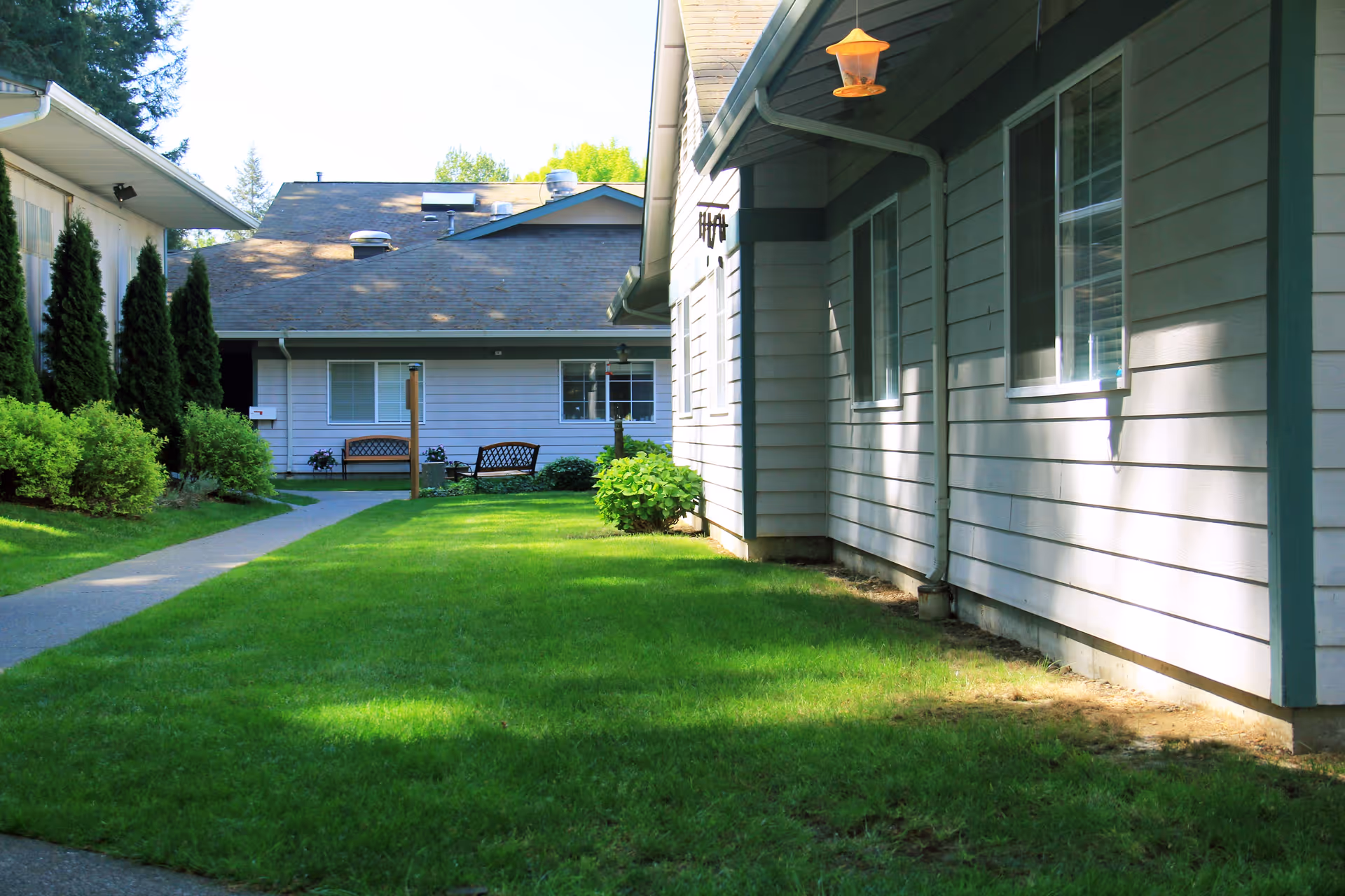 Outdoor view of a senior living facility with green grass, a concrete pathway, bushes, and two benches near the building. The buildings have light-colored siding and multiple windows, with trees visible in the background under a clear sky.