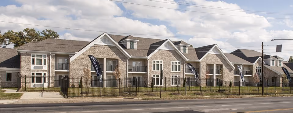 Exterior view of a large, modern senior living facility building with stone facade, multiple windows, balconies, and a fenced lawn. Several black flags with 'NOW OPEN' text are placed along the front of the building. The sky is partly cloudy.