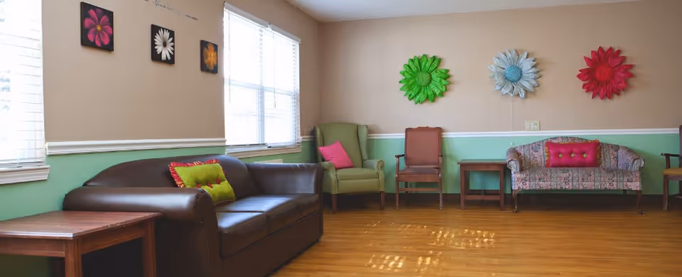 A cozy sitting area in a senior living facility with a brown leather sofa, a green armchair, a floral patterned loveseat, and wooden chairs. The walls are painted beige and green with colorful flower decorations and framed flower pictures. Large windows allow natural light to fill the room.