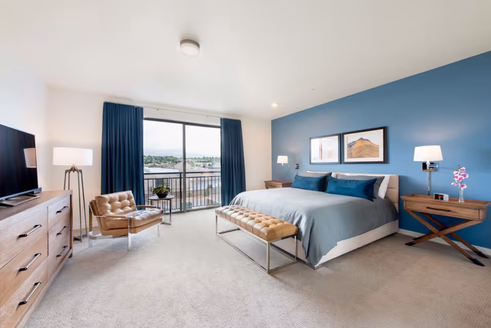 A modern bedroom with a large bed featuring blue bedding and pillows against a blue accent wall. Two framed pictures hang above the bed. There are two wooden nightstands with lamps on either side of the bed, one with a small vase of pink flowers. A tufted leather bench sits at the foot of the bed. A leather armchair and a wooden dresser with a flat-screen TV are positioned near a large window with blue curtains, offering a view of buildings outside.