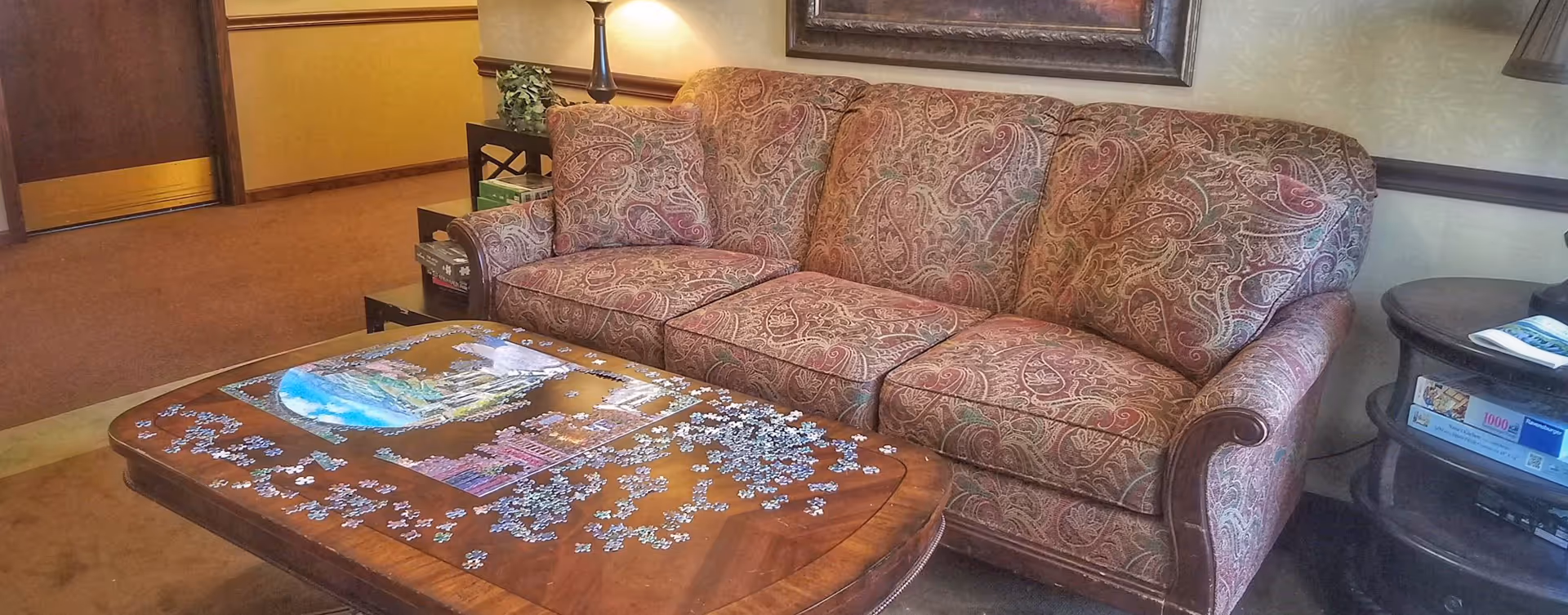 Upholstered patterned sofa next to a wooden coffee table strewn with jigsaw puzzle pieces in a cozy sitting area.