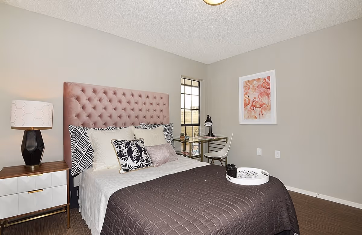 A neatly made bed with a tufted pink headboard, white and patterned pillows, and a dark brown quilt. Next to the bed is a modern nightstand with a geometric black and white lamp. A small desk with a white chair and a black desk lamp is positioned near a window with black framing. A framed artwork with pink flamingos hangs on the beige wall.