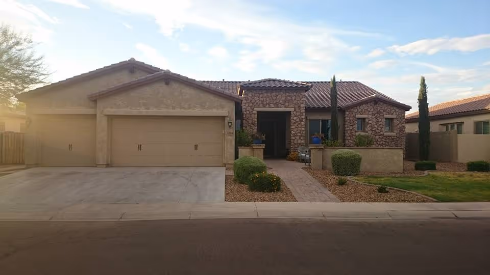 Front exterior view of a single-story house with a three-car garage, stone and stucco facade, a tiled roof, and a landscaped front yard with bushes, small trees, and a paved walkway leading to the entrance.