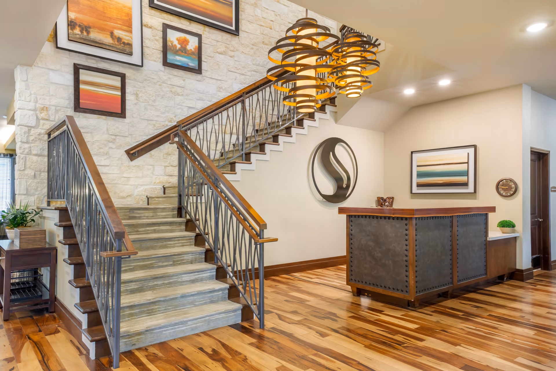 Interior view of a senior living facility lobby featuring a wooden staircase with metal railings, a reception desk with a metal front and wooden top, modern pendant lighting, framed artwork on the walls, and a polished wooden floor.