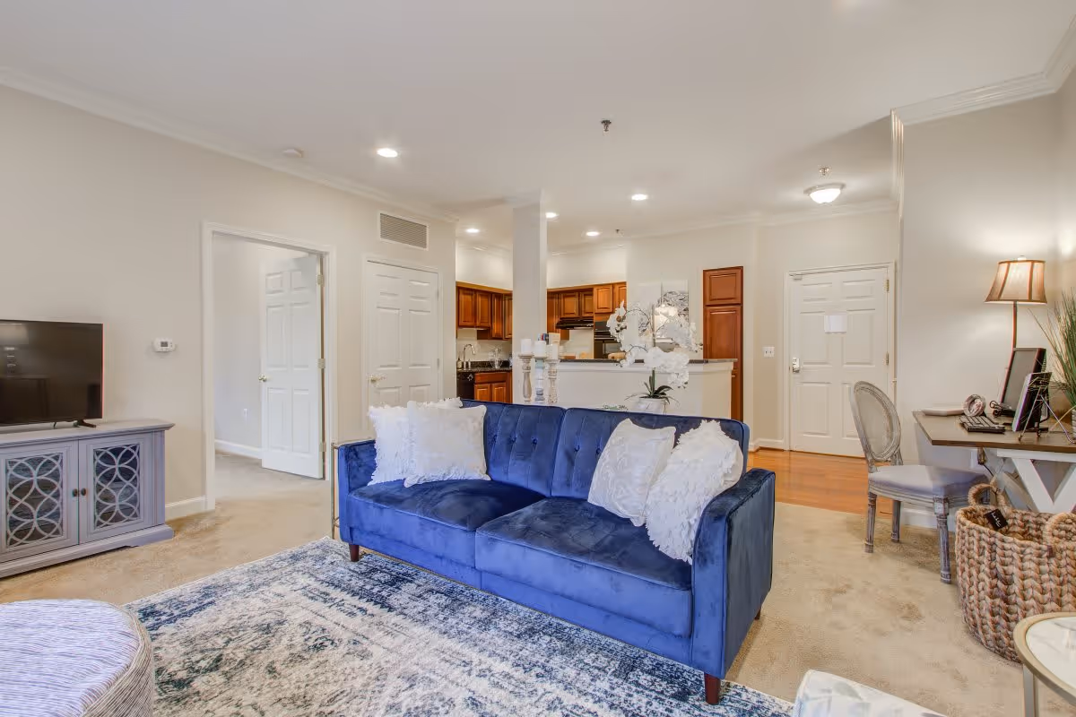 A bright and spacious living room with a blue velvet sofa adorned with white pillows, a patterned area rug, and a TV on a gray cabinet. The room opens into a kitchen with wooden cabinets and a countertop. There is a small desk with a chair and a lamp on the right side near the entrance door. The walls are painted light beige, and the floor is carpeted except for the kitchen area which has wooden flooring.