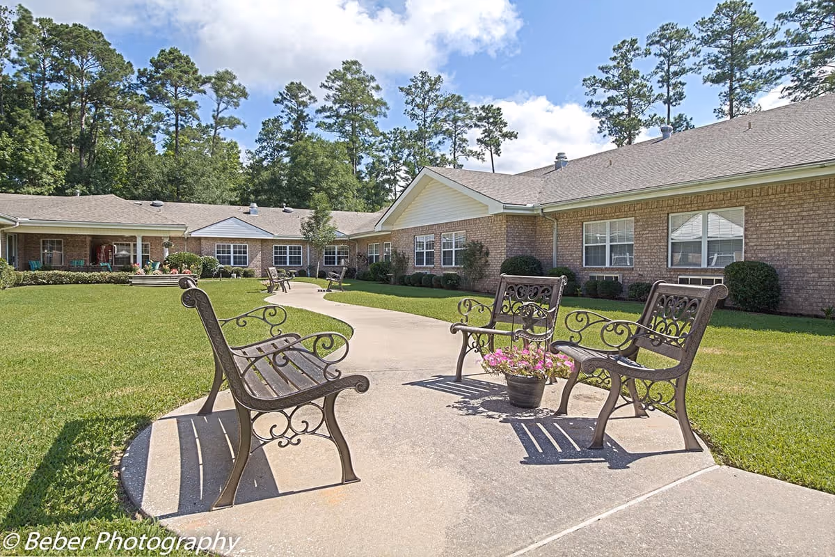 Outdoor courtyard with decorative metal benches and a paved walkway in front of a single-story brick senior living building.