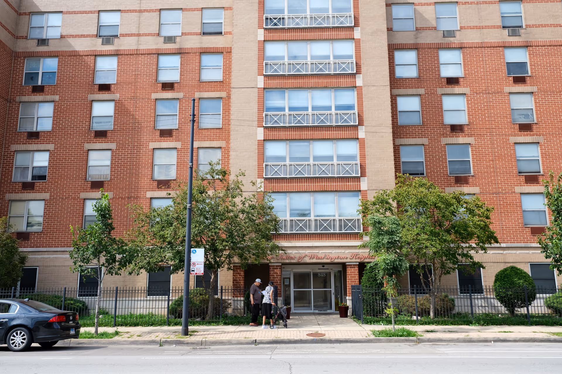Exterior front of a multi-story brick apartment building with a central entrance, trees, and two people standing on the sidewalk.