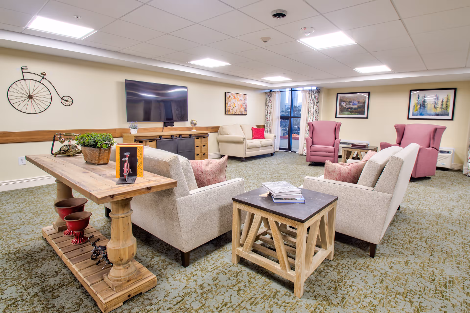 A cozy living room area in a senior living facility with beige armchairs and sofas, two pink wingback chairs, a wooden coffee table with books, a wooden console table with decorative items, a wall-mounted flat screen TV, and framed artwork on the walls. The room has carpeted flooring and large windows with floral curtains.