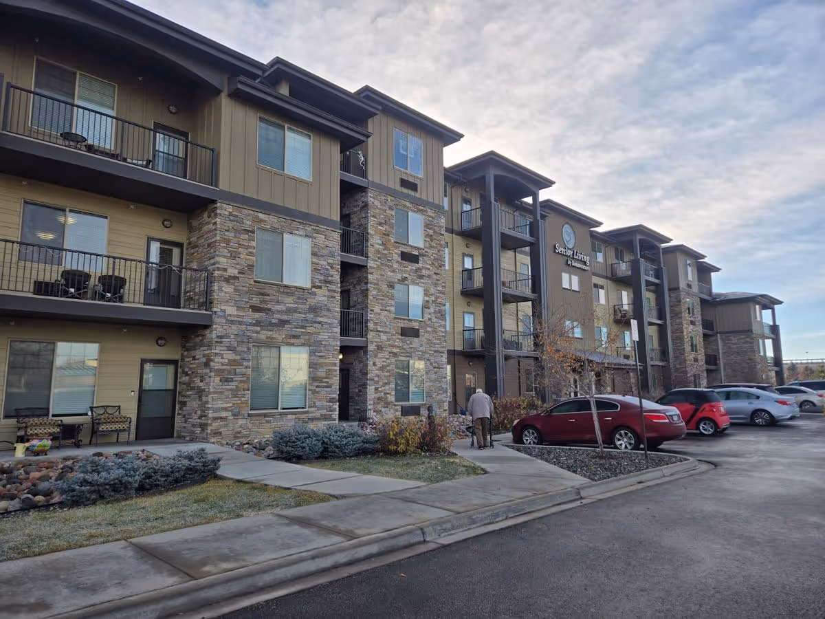 Front exterior of a multi-story senior living building with balconies, parked cars, and a person walking toward the entrance.