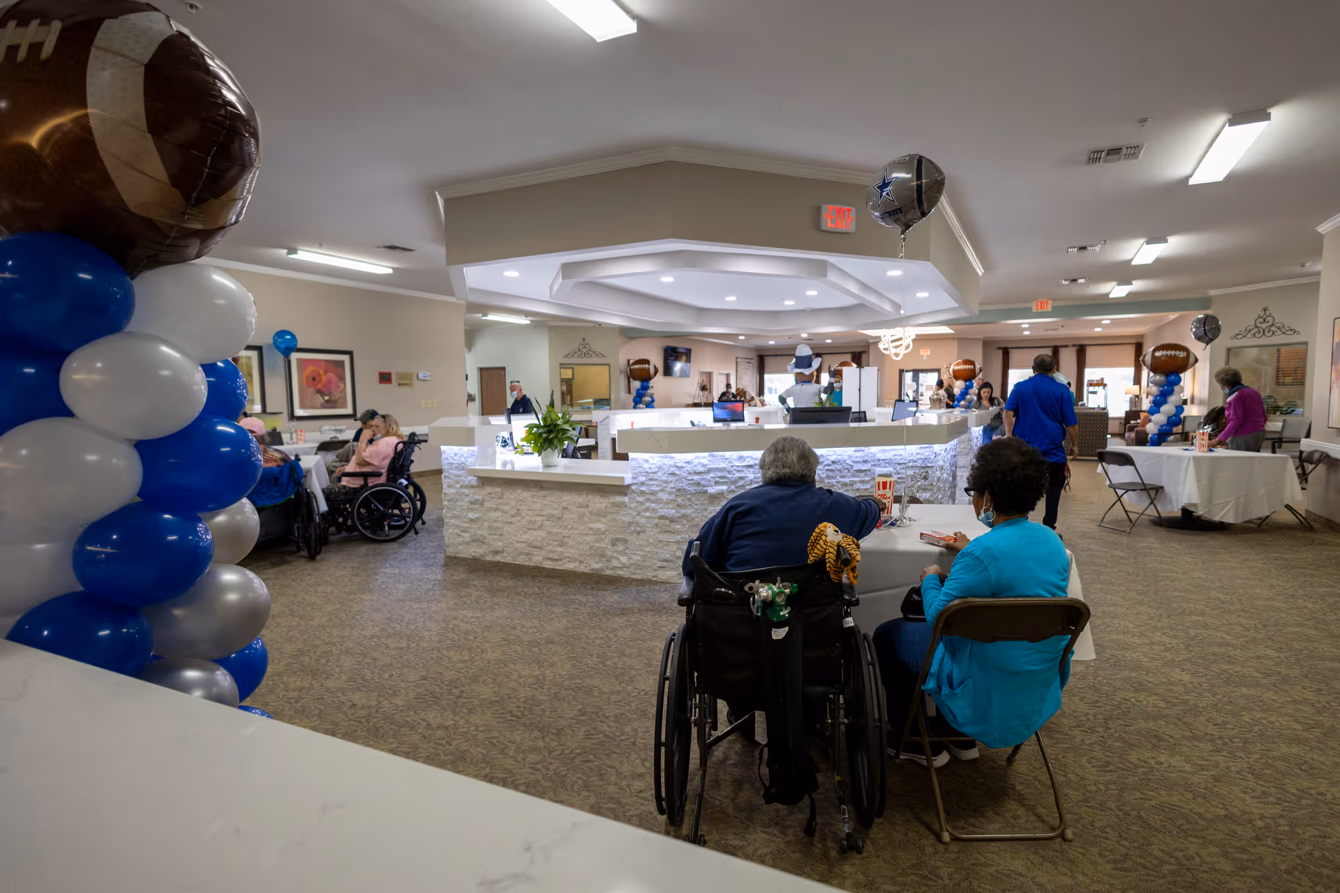 Communal dining/lounge area in a senior living facility with residents seated at tables, balloons, and a central reception desk.