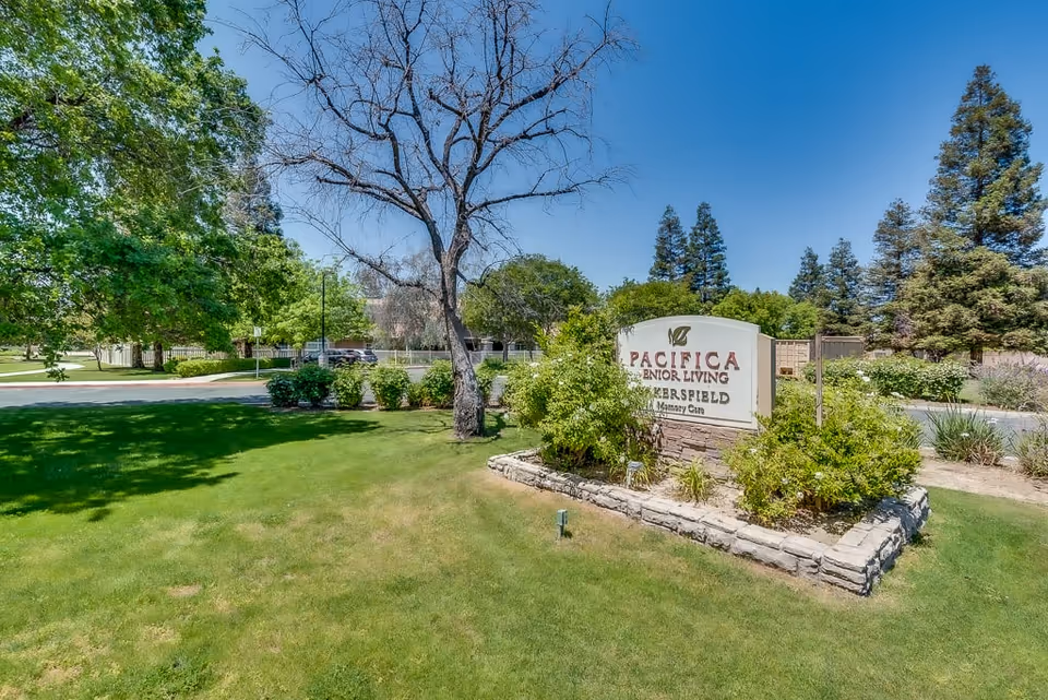 Outdoor view of a senior living facility sign that reads 'Pacifica Senior Living Bakersfield Memory Care' surrounded by green grass, bushes, and trees under a clear blue sky.