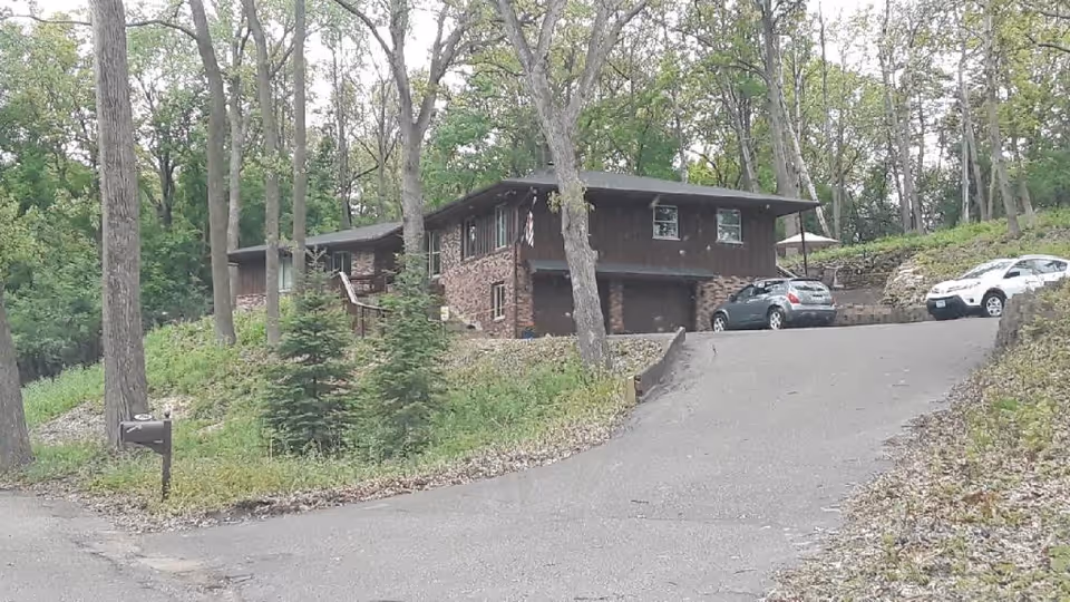 Brick-and-wood split-level house surrounded by trees with a sloped driveway, mailbox, and two cars parked near the garage.