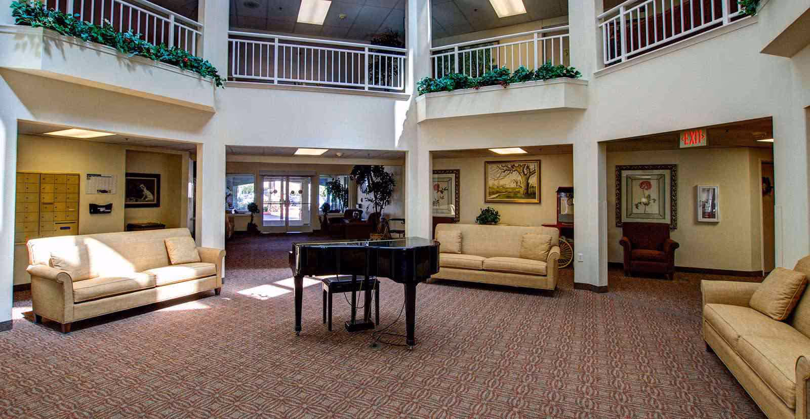 Spacious senior living facility common area with three beige sofas arranged around a black grand piano. The room has a patterned carpet, white walls, and an upper balcony with railings decorated with green plants. There are framed pictures on the walls and an exit sign above a doorway.