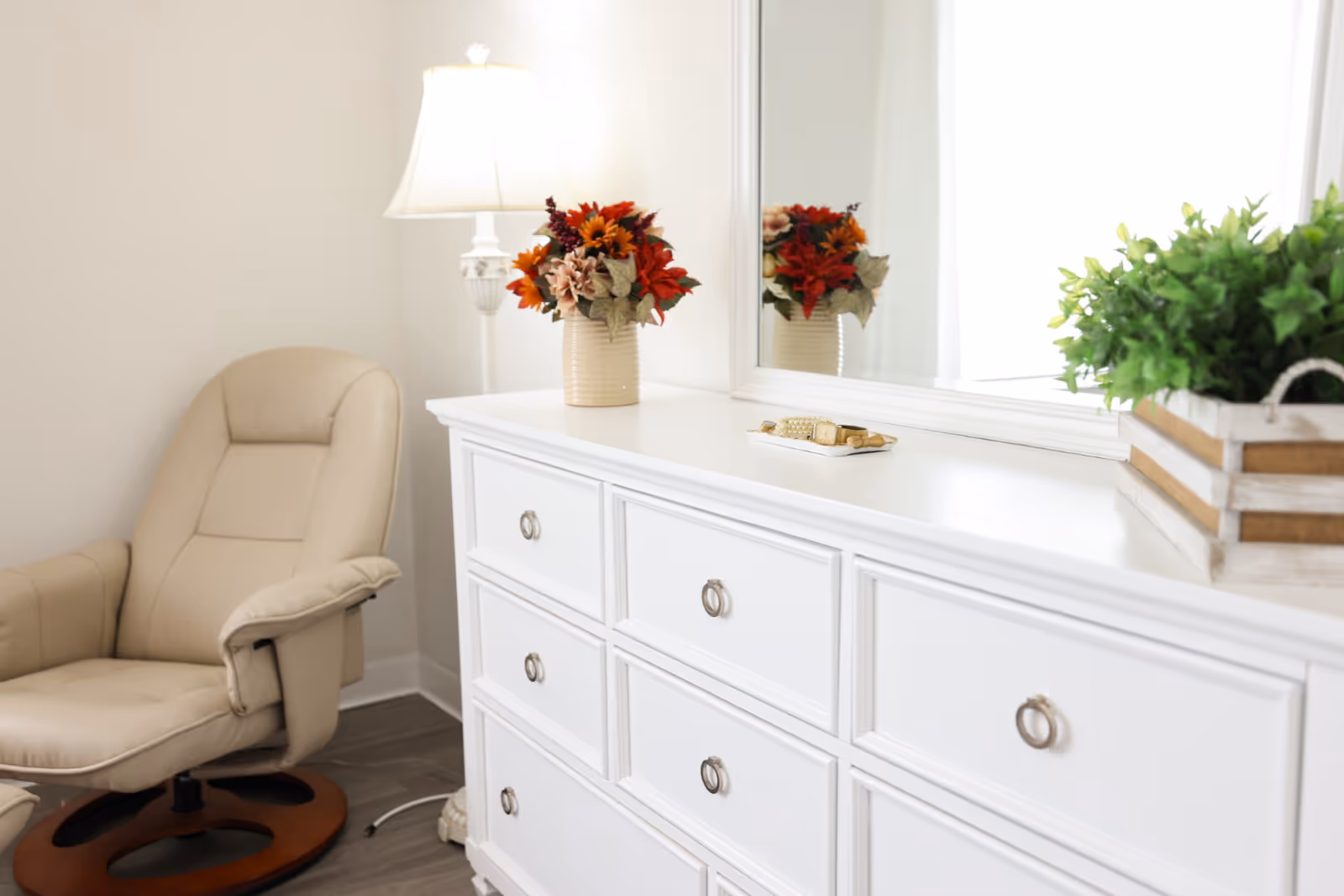 A cozy corner of a room featuring a beige cushioned armchair with a wooden base, a white dresser with multiple drawers and round metal handles, a large mirror above the dresser, a white floor lamp with a white lampshade, a vase with autumn-colored flowers, and a small wooden crate with green plants on top of the dresser.