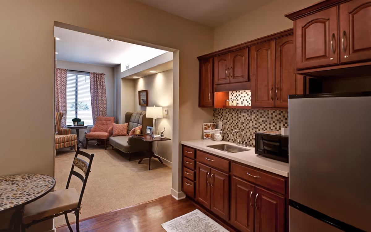 Interior view of a senior living facility apartment showing a small kitchen area with wooden cabinets, a sink, a microwave, and a refrigerator. Adjacent to the kitchen is a cozy living room with a sofa, two armchairs, a side table with a lamp, and a window with patterned curtains letting in natural light.