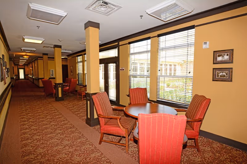 A long hallway in Webb House Retirement Center with patterned carpet and beige walls. Along the right side, there are large windows with blinds and a round wooden table surrounded by four red upholstered chairs. The hallway has several columns and framed pictures on the walls, with additional seating areas visible further down the corridor.