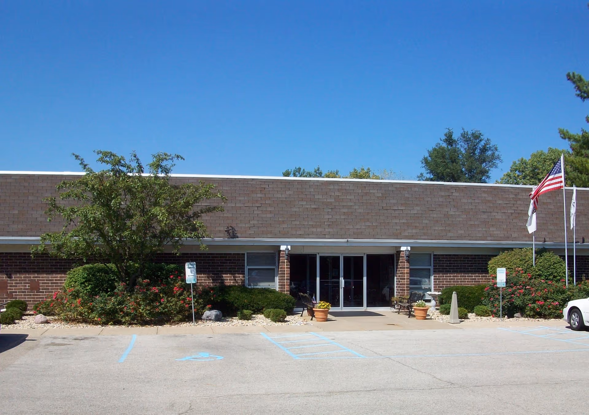 Exterior front view of a single-story brick building with a flat roof, featuring a main entrance with glass double doors. There are two flagpoles with flags on the right side, a tree and bushes in front of the building, and a parking lot with handicap parking spaces in the foreground under a clear blue sky.