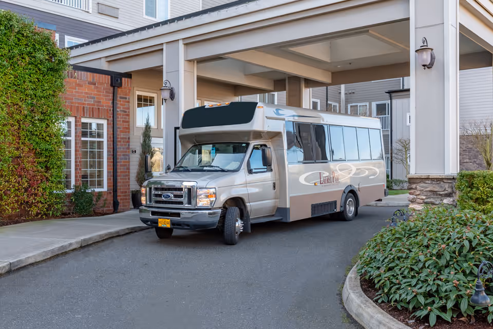 A beige shuttle bus parked under a covered entrance of a senior living community building. The building features brick and siding exterior with windows and greenery around the driveway.