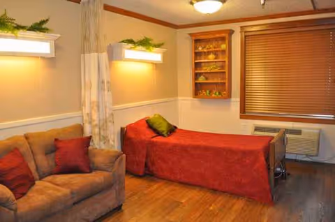 Cozy senior living room with beige sofa, red-covered bed, wooden floor, and window blinds.