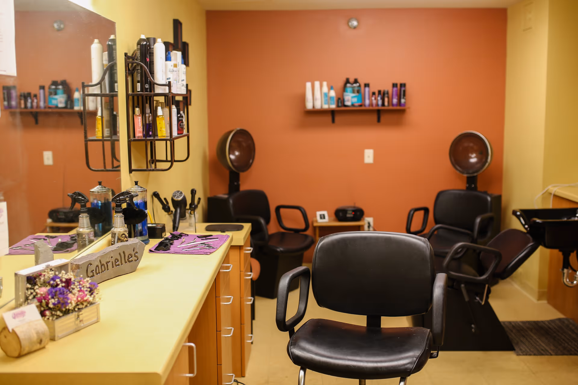 Interior view of a hair salon area with a large mirror, a countertop holding various hair care products and tools, a sign that reads 'Gabrielle's', black salon chairs, hair dryers, and shelves with additional hair products against a warm-toned wall.