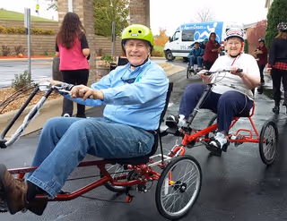 Two elderly individuals wearing helmets are riding red recumbent tricycles on a paved surface outside a building entrance. Other people are visible in the background near a white van with blue graphics.