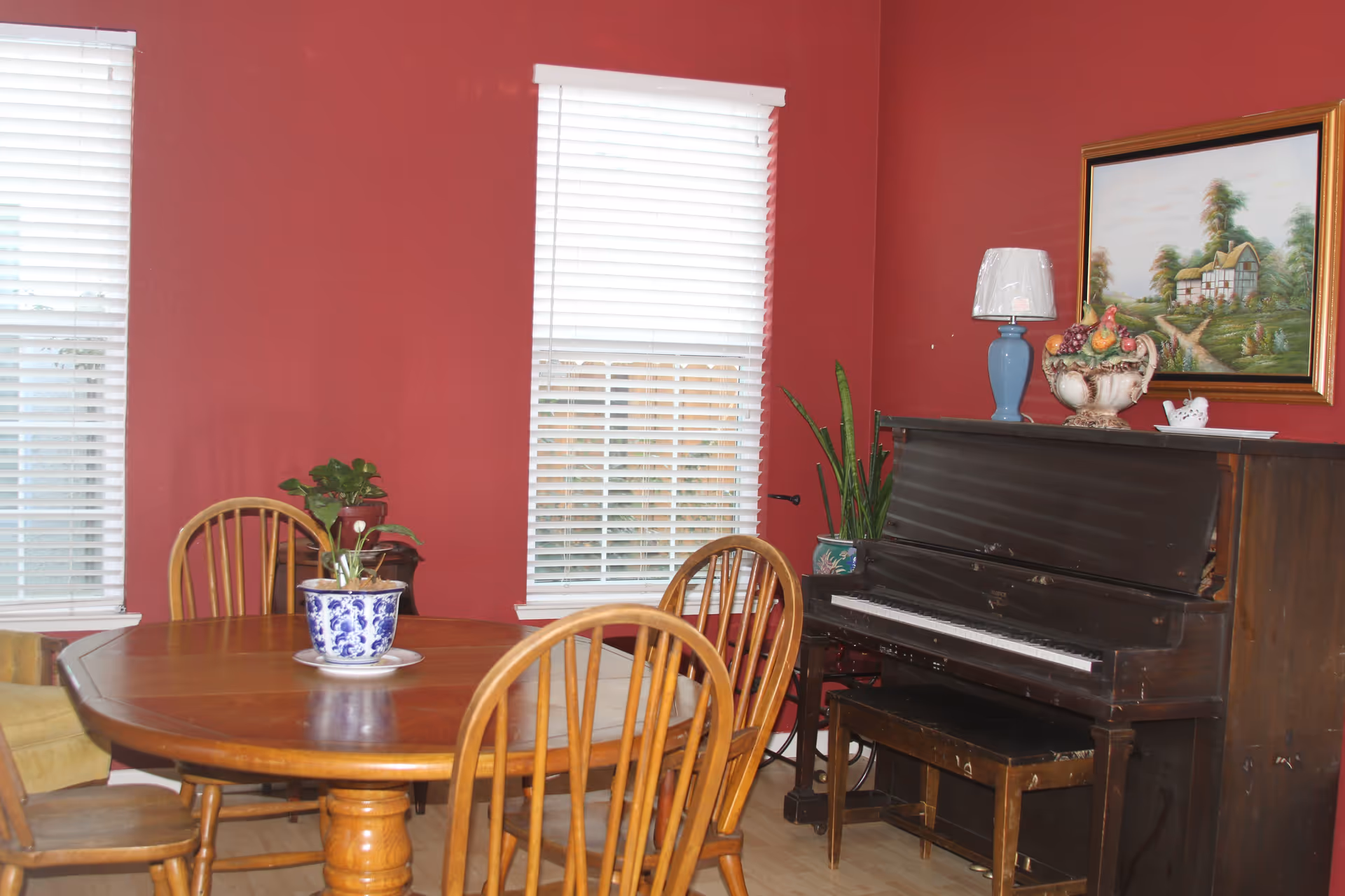 A cozy room with red walls featuring a wooden dining table surrounded by wooden chairs. There are two windows with white blinds behind the table. To the right, there is an upright piano with a bench, decorated with a blue lamp, a fruit basket, and a potted plant. A framed painting of a countryside house hangs above the piano.