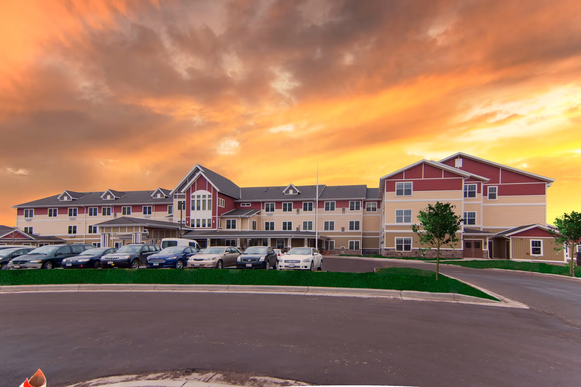 Front exterior of a multi-story senior living facility with parked cars and a dramatic sunset sky.