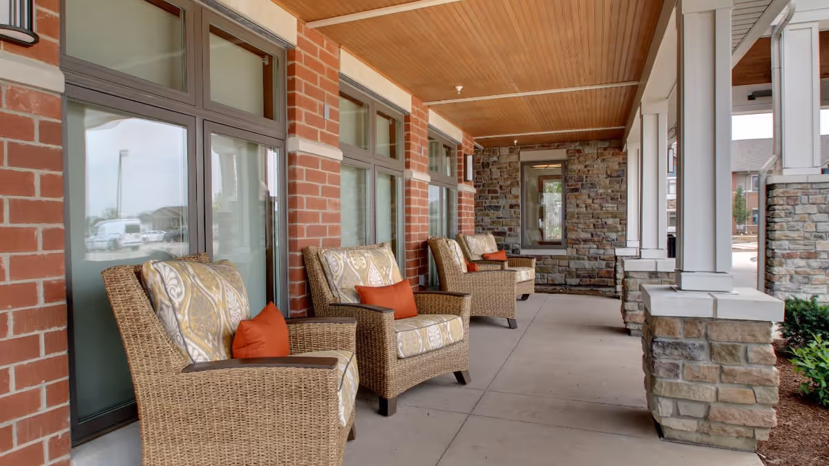 Covered outdoor patio area with wicker armchairs featuring patterned cushions and orange throw pillows, brick and stone walls, and large windows along one side.