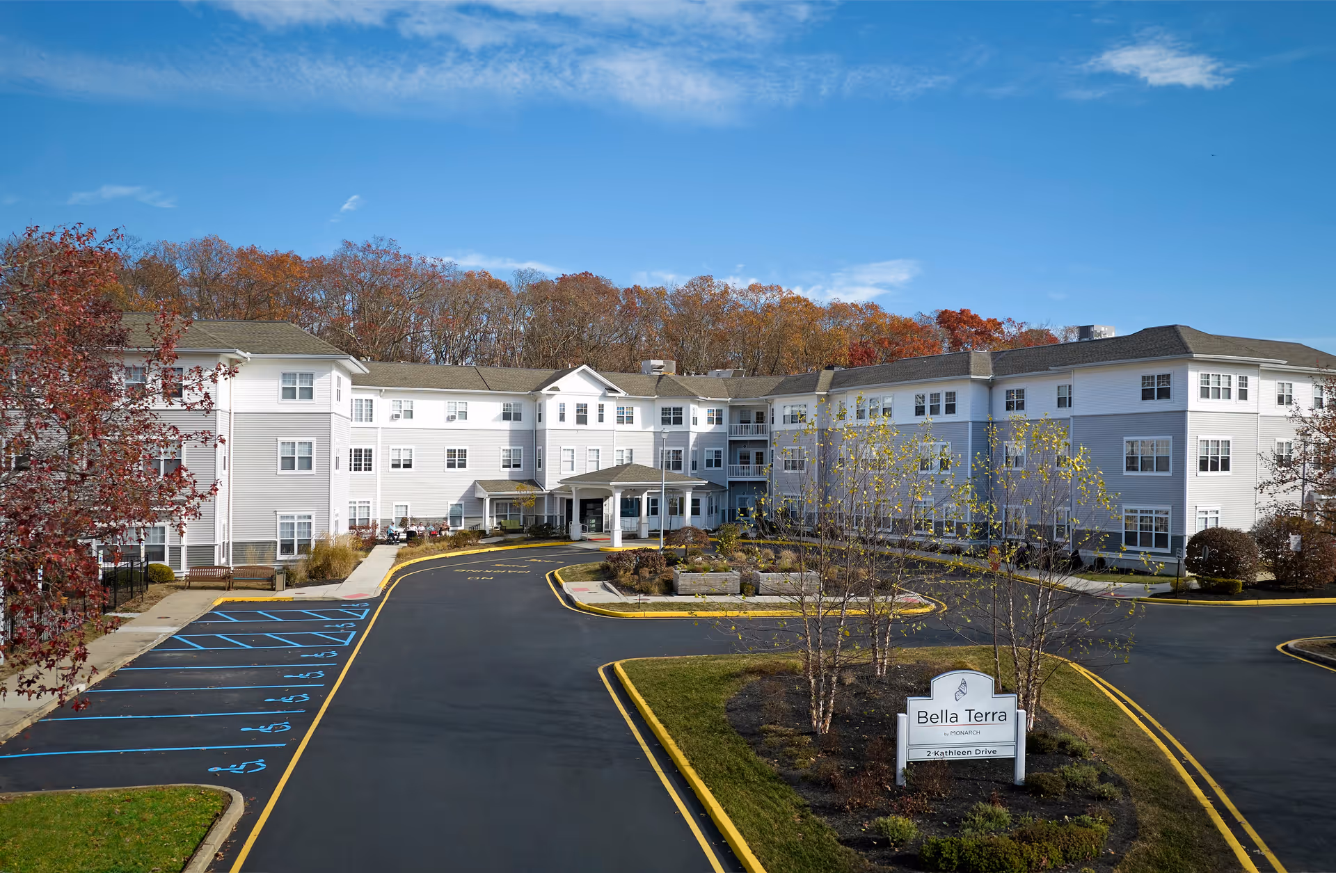 Exterior view of Monarch Bella Terra, a three-story senior living facility with a large parking area featuring several handicapped parking spaces. The building is surrounded by trees with autumn foliage under a clear blue sky. A sign in front reads 'Bella Terra by Monarch, 2 Kathleen Drive.'
