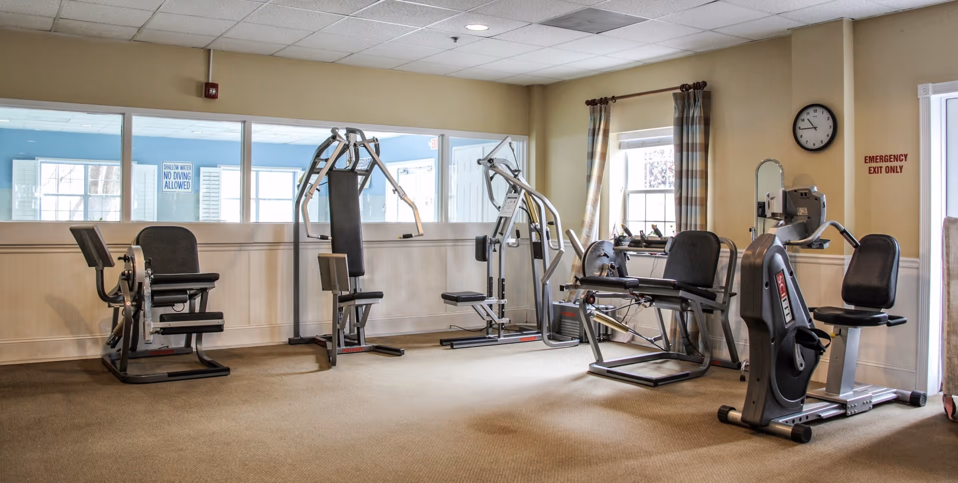 A fitness room with various exercise machines including a leg press, a multi-gym machine, and stationary bikes. The room has beige walls, a carpeted floor, a window with plaid curtains, a clock on the wall, and an emergency exit door.