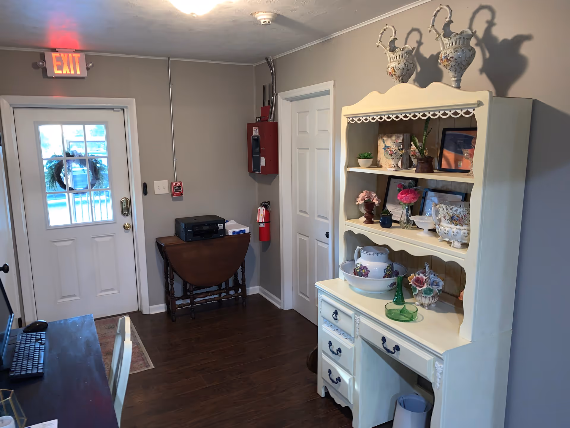 Interior room with a white door featuring a window and a wreath, an exit sign above the door, a small wooden table with a printer and paper on it, a fire extinguisher and fire alarm on the wall, and a cream-colored hutch displaying decorative vases, flowers, and framed pictures. The room has wooden flooring and light gray walls.
