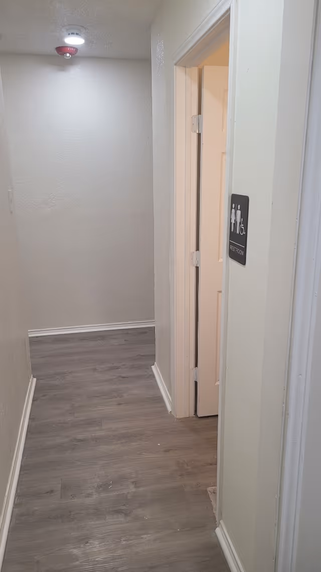 Narrow interior hallway with wood-look flooring leading to an open restroom door marked with an accessible restroom sign.