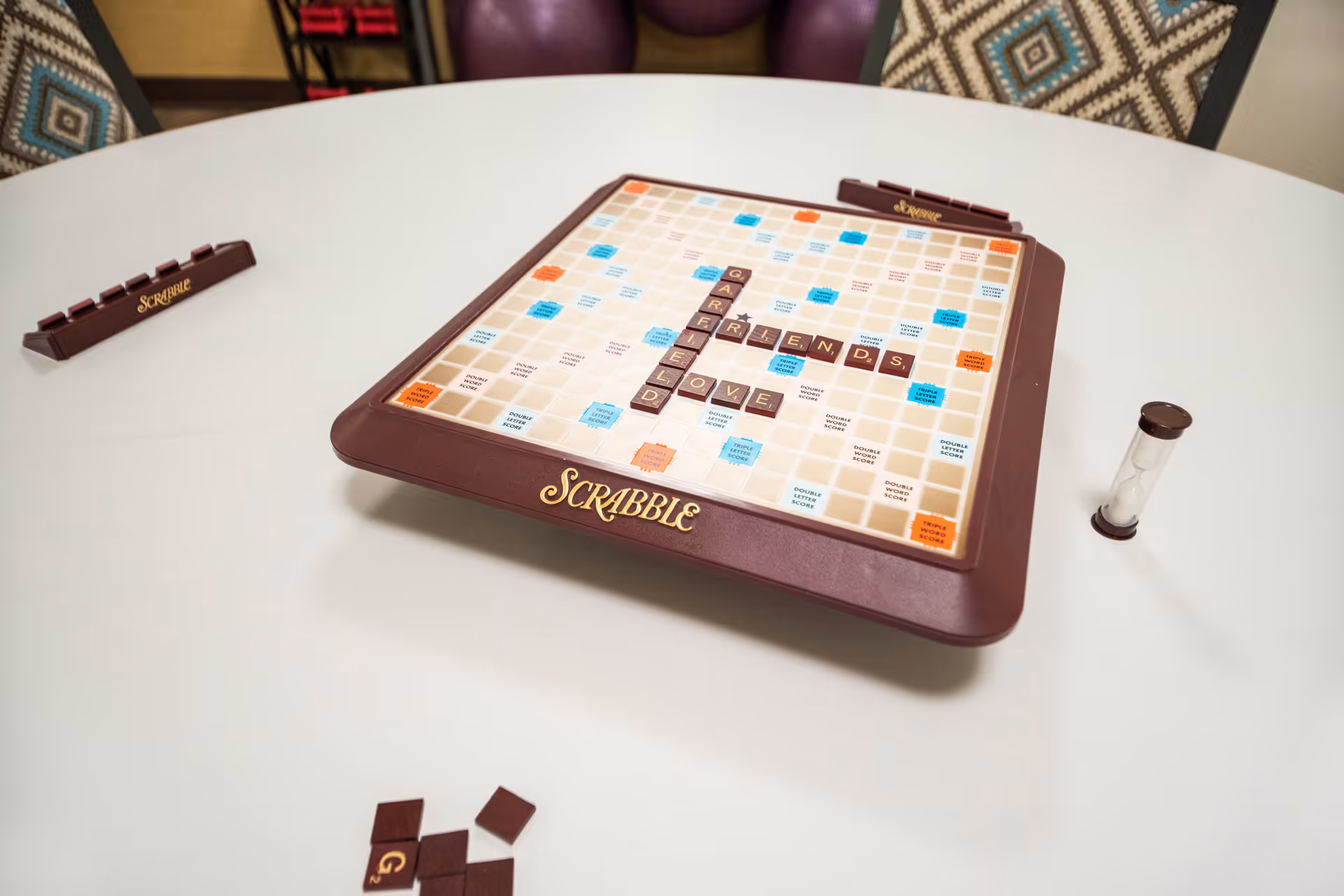 A Scrabble board on a white round table with the words GARFIELD, FRIENDS, and LOVE spelled out. There are Scrabble tile racks, some loose tiles, and a sand timer on the table. In the background, there are patterned cushions on chairs.