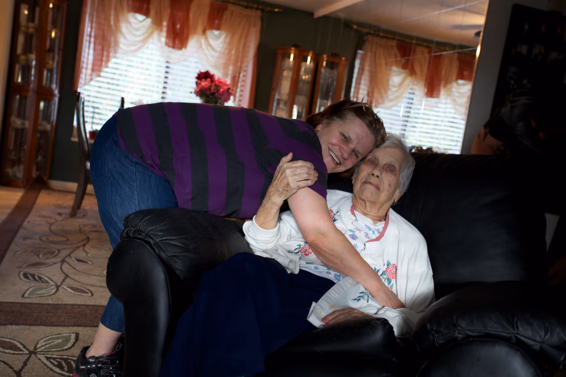 An elderly woman sitting on a black leather armchair with a younger woman leaning over and hugging her in a warmly lit living room with large windows covered by sheer curtains and a floral carpet on the floor.
