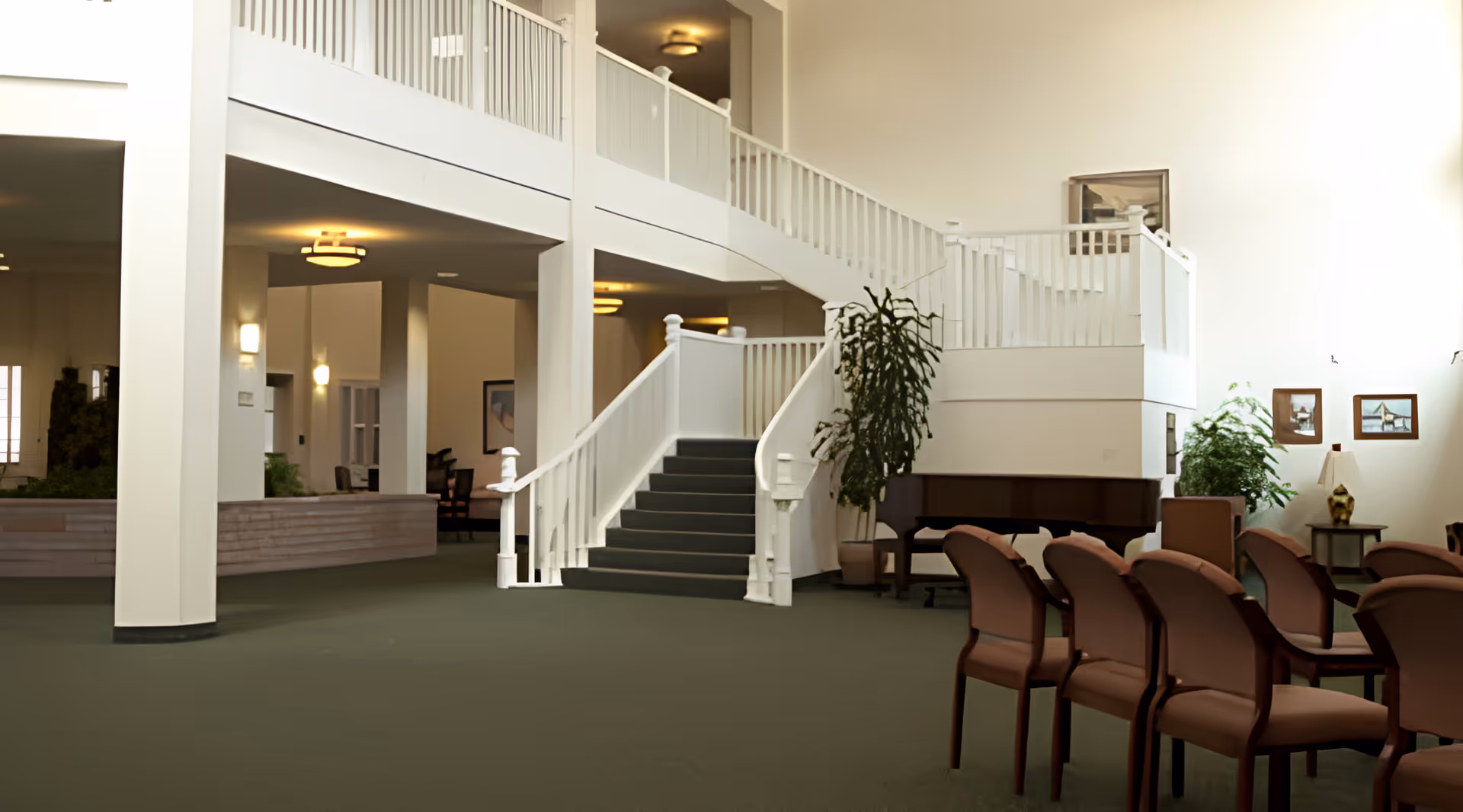 Spacious interior of a senior living facility with a green carpeted floor, a white staircase with railings leading to an upper level, several brown chairs arranged in rows, a grand piano, potted plants, and framed artwork on the walls.