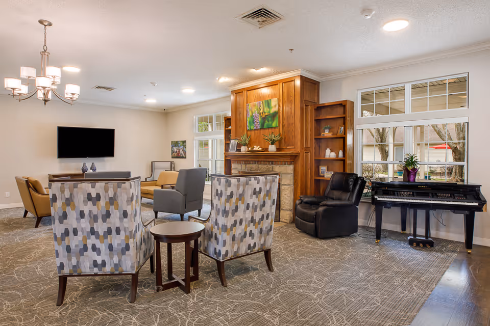 A spacious communal living room with patterned armchairs around a small table, a fireplace with wooden shelving, a wall-mounted TV, and a piano by the window.