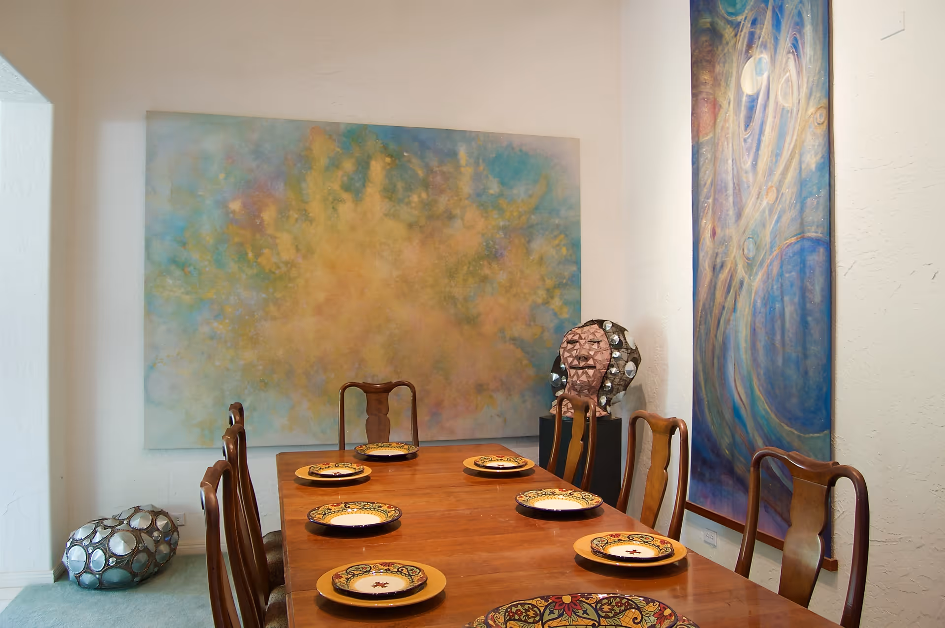 A dining room with a wooden table set with decorative plates and six wooden chairs. The walls feature two large abstract paintings, one with warm yellow and blue tones and the other with swirling blue and purple patterns. There are also two decorative sculptures placed near the walls.